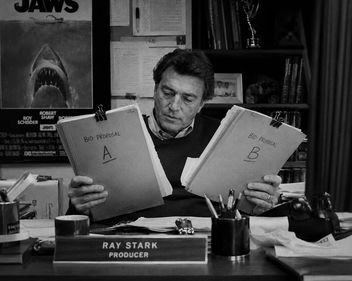 A black-and-white 1980s production office still with a producer comparing two bid proposal folders.