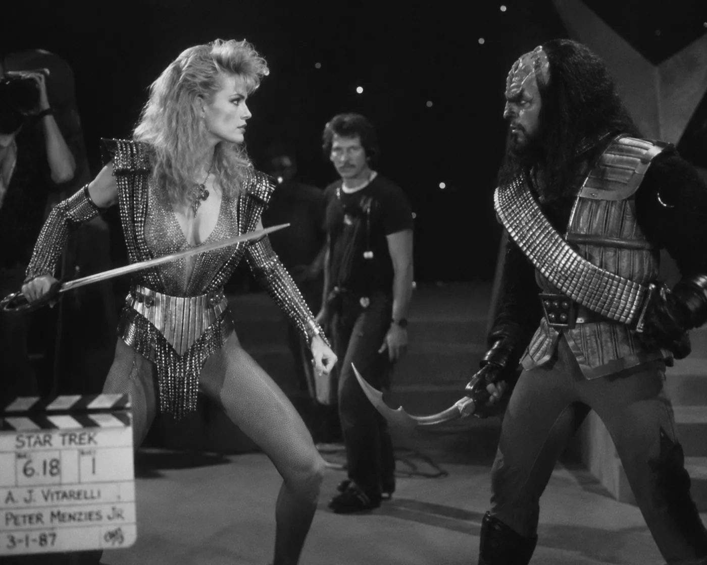 A black-and-white 1980s film set still with two costumed performers facing off while crew watch nearby.