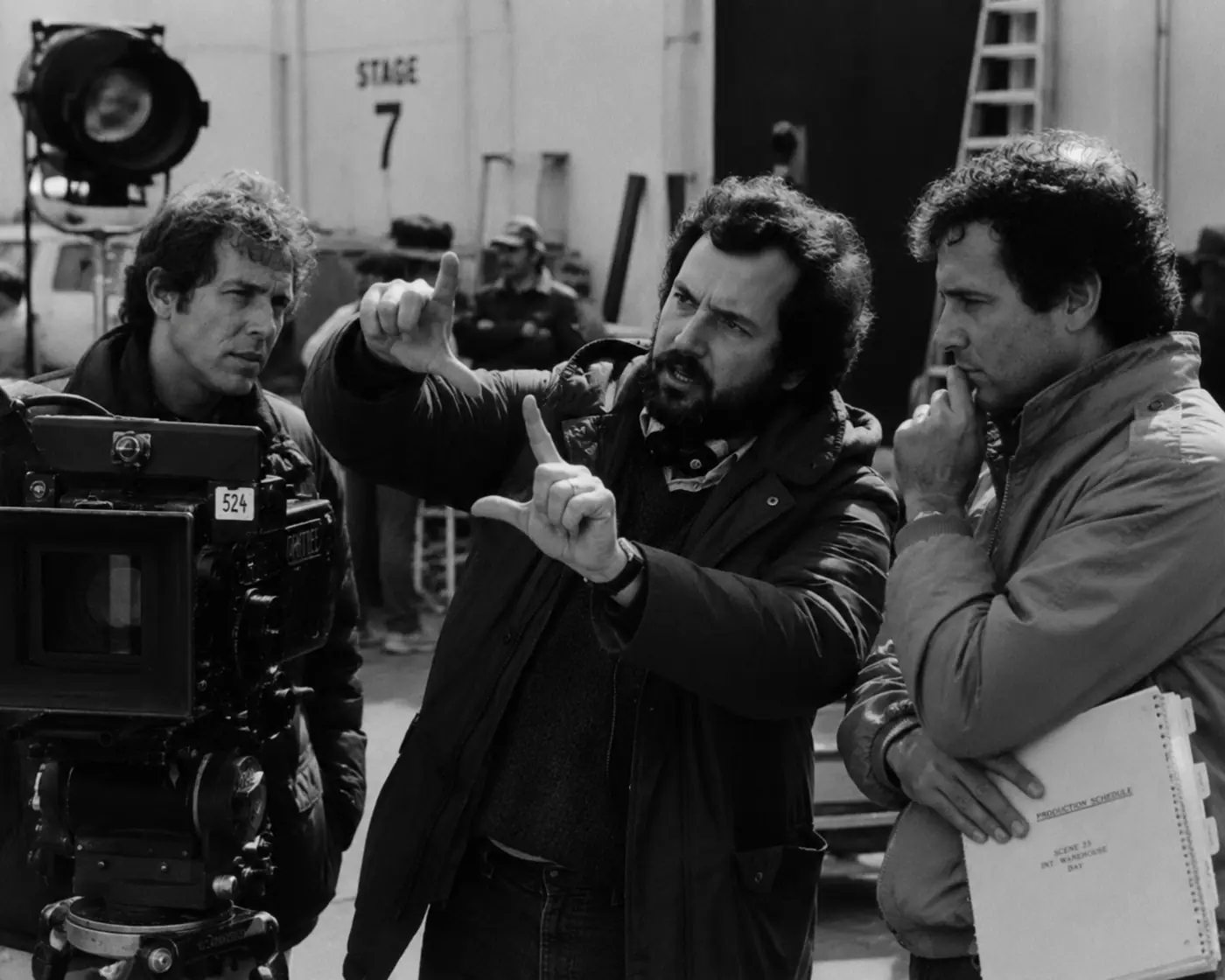 A black-and-white 1980s film crew plans a shot with frame photos, storyboards, lenses, a CRT monitor, and a 35mm camera.